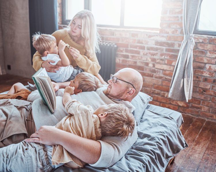 Joyful father and mother playing with their children in living room and teaching them to read a book with fairytale.